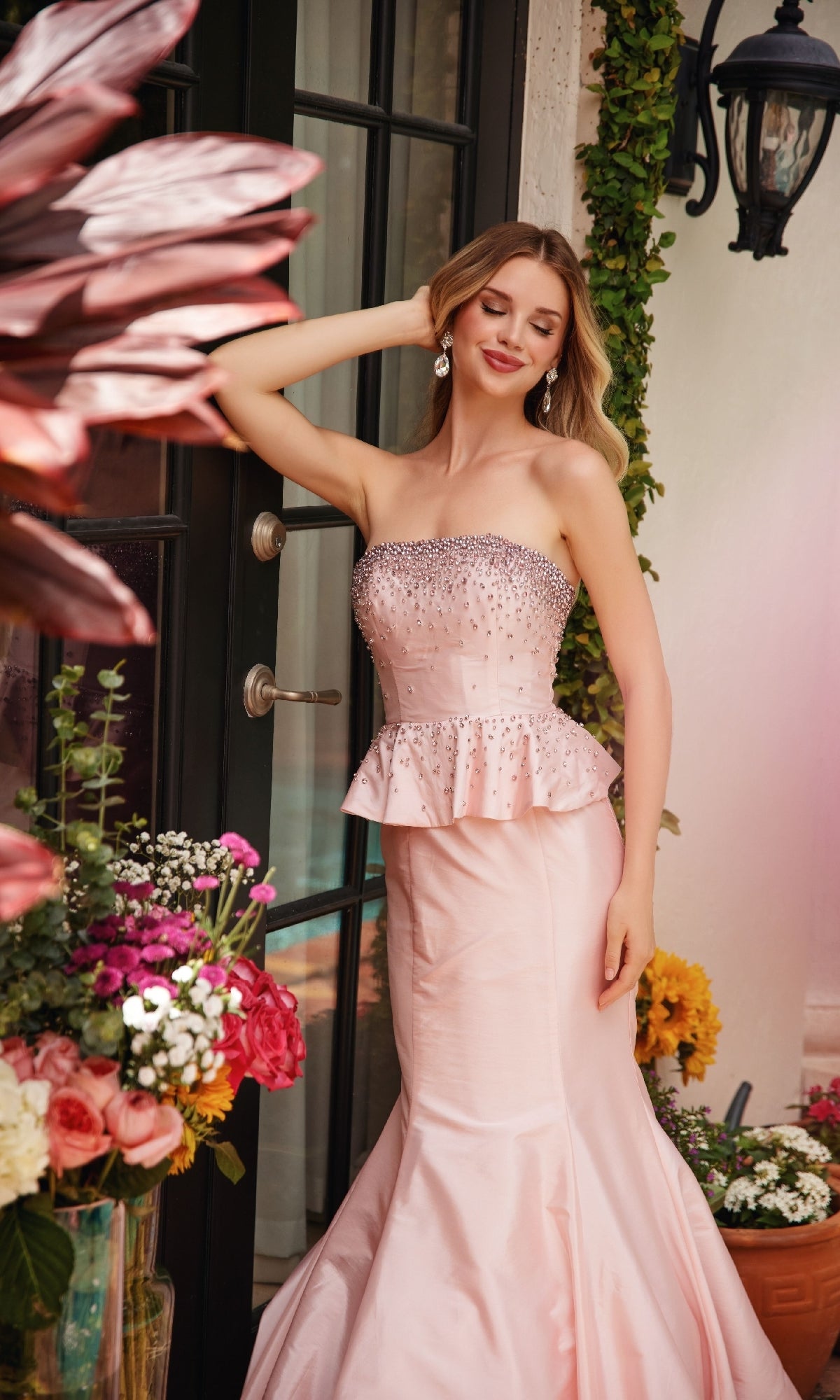 A woman in the Ava Presley 40092 Strapless Peplum Prom Dress, featuring rhinestone embellishments and a mermaid silhouette, stands by a glass door surrounded by potted flowers.
